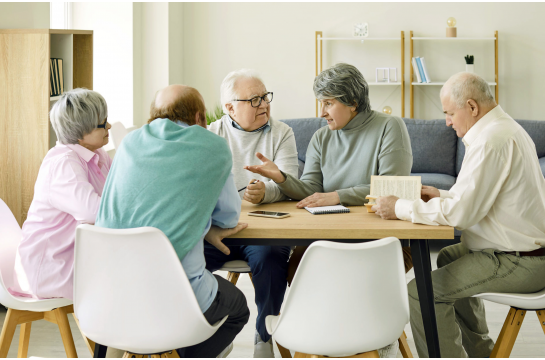 Fotografie: Fünf Seniorinnen und Senioren sitzen gemeinsam mit einem Buch am Tisch und reden.