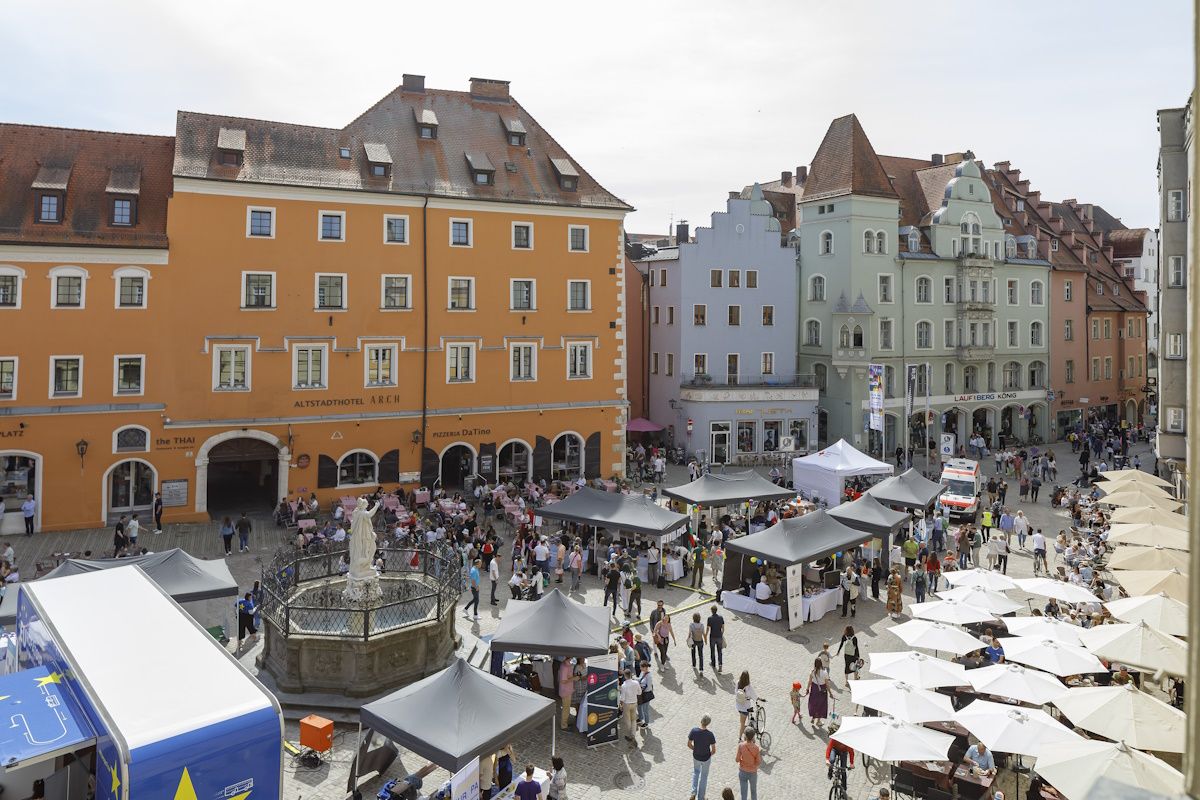 Blick von Norden von oben auf den Regensburger Haidplatz. Viele verschiedene Stände sind gut besucht und ein blauer Bus mit Werbung für Europa steht östlich vom Brunnen.