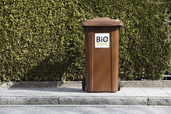 Fotografie: Eine braune Biotonne steht vor einer Hecke auf dem Gehweg.