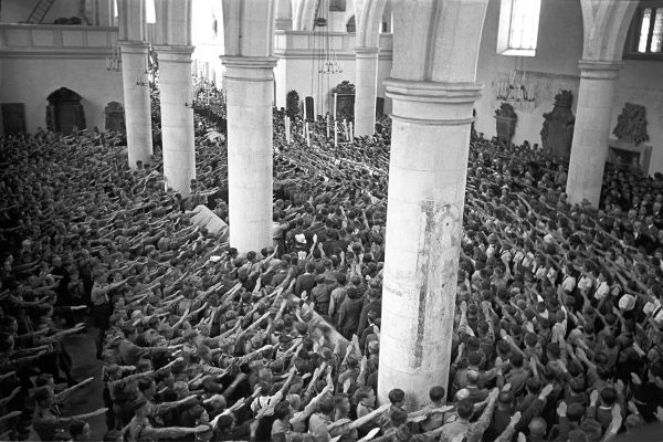 Zu sehen ist eine Versammlung in der Zeit des Nationalsozialismus. Die Versammlung findet in einer Kirche statt. Es sind sehr viele Menschen in der Kirche zu sehen. Alle zeigen einen Hitlergruß in Richtung Altar.  