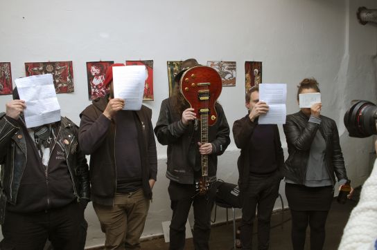 Mitglieder der Schreibwerkstatt Salamander stehen vor einer Wand mit ausgestellten Bildern, die Gesichter verdeckt durch hochgehaltene bedruckte Blätter und eine E-Gitarre. (C) Wolfgang Ruhl