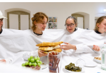 Fotografie: Vier Personen stecken in einem großen weißen Laken mit Öffnungen für Hände und Köpfe und reichen einen Teller mit Fladenbrot durch.