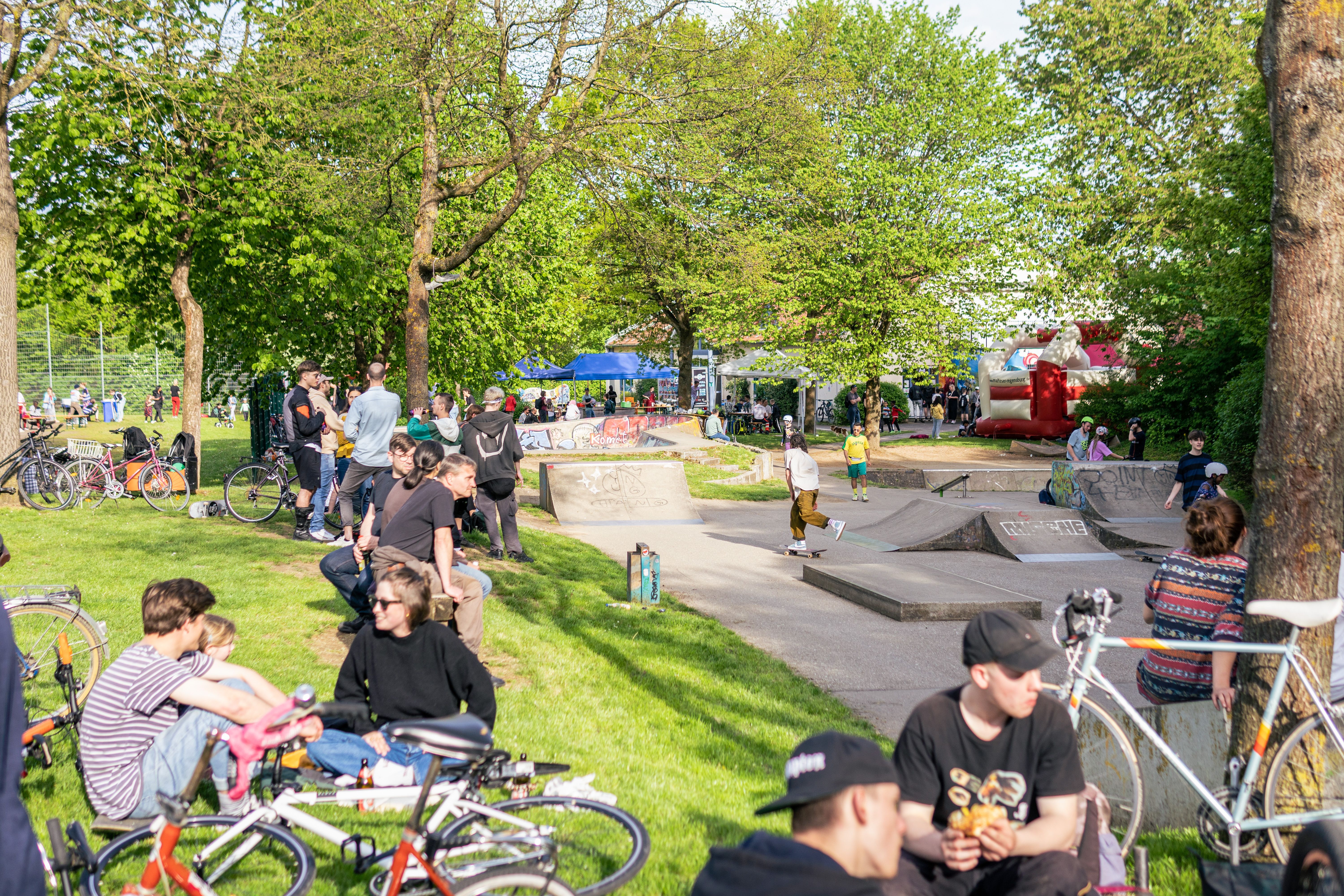 viele Menschen genießen den sonnigen Tag auf dem Skatepark. In der Mitte ist ein Skateboarder zu sehen, an den Bildrändern diverse Fahrräder
