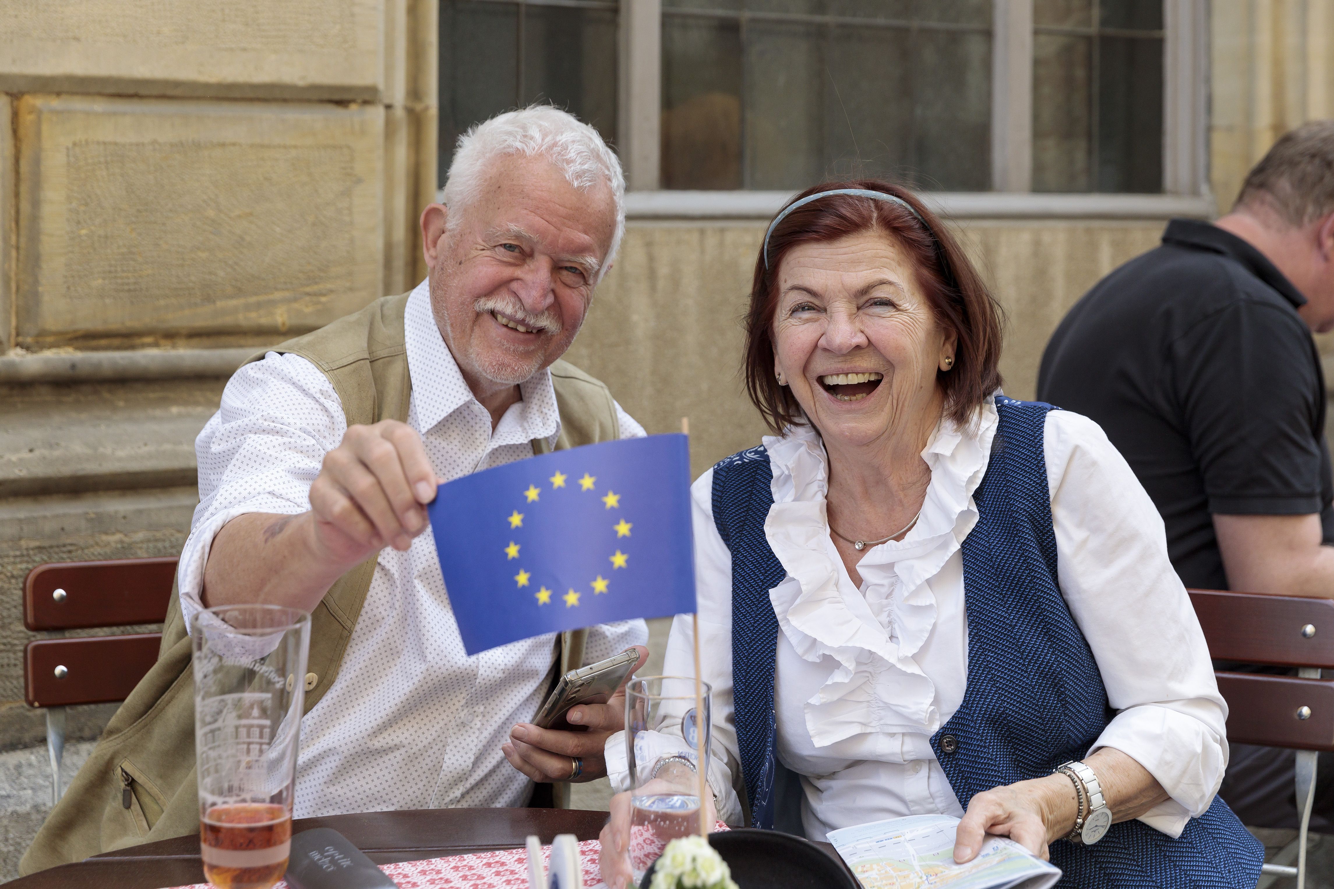 älteres Ehepaar sitzt an einem runden Biergartentisch. Der Mann zeigt eine kleine Europa-Flagge in die Kamera.