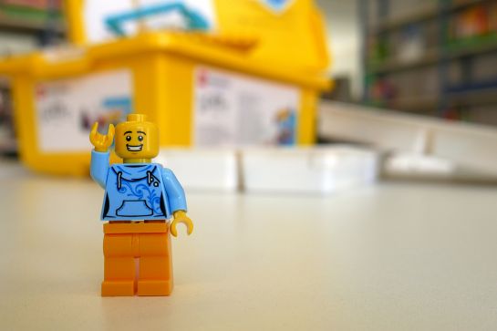 Legofigur Spike winkend. Dahinter Box mit Lego, unscharf zu erkennen