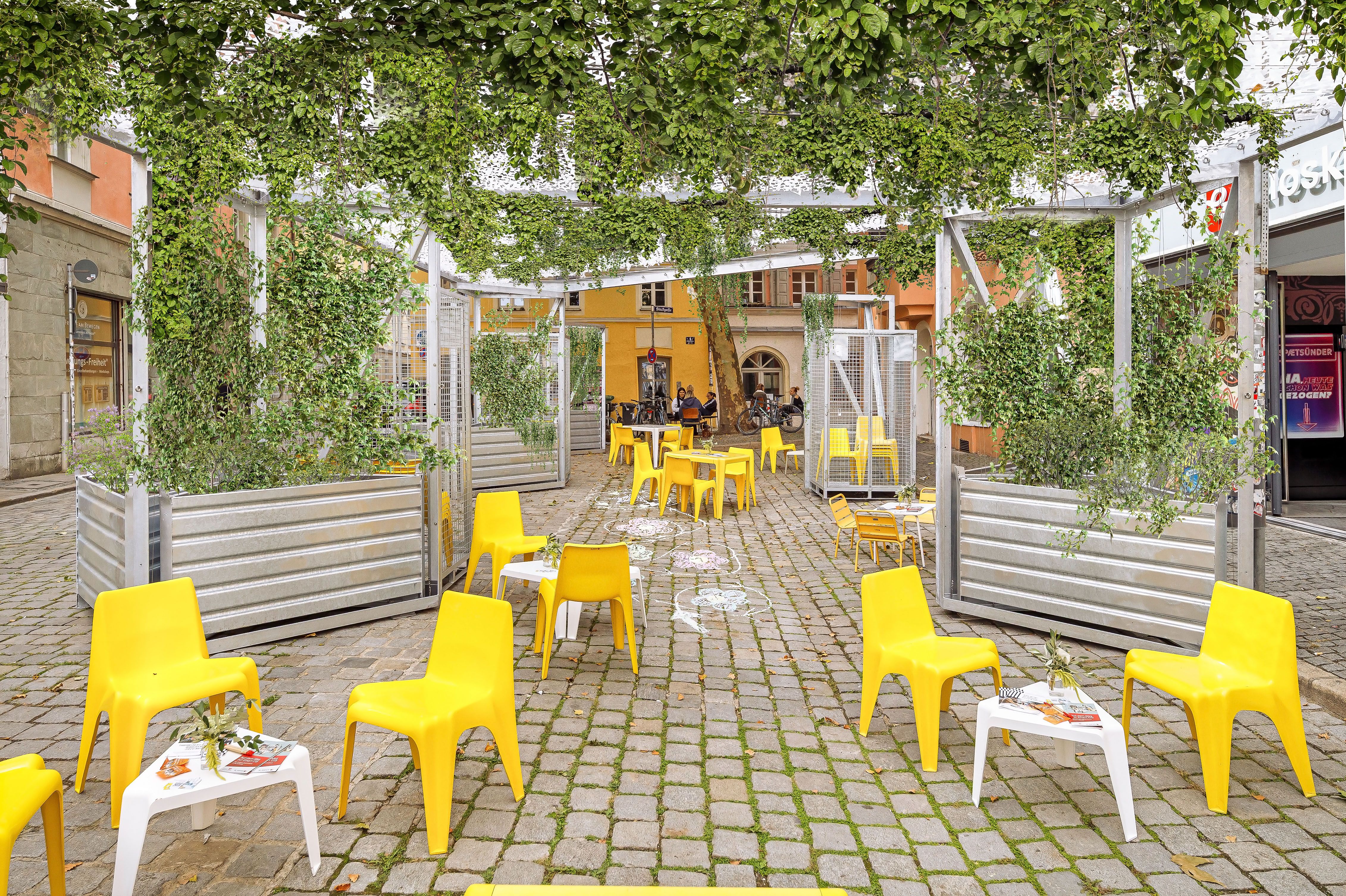 Fotografie – Visualisierung der neuen grünen Oase in der Obermünsterstraße: Tische und Stühle unter den begrünten Stahlkonstruktionen