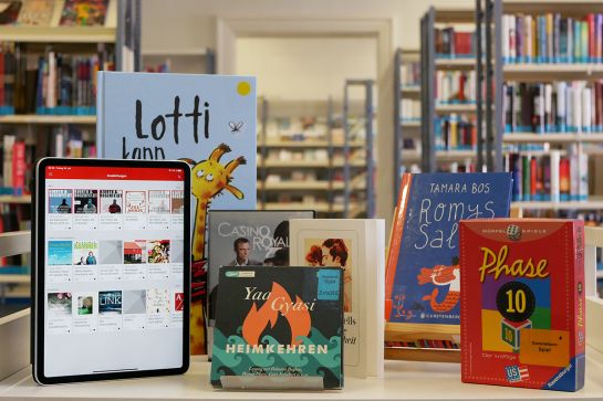 Tisch auf dem Bücher, Spiel, DVD, CD stehen, im Hintergrund Büchereiregale (C) Andrea Borowski