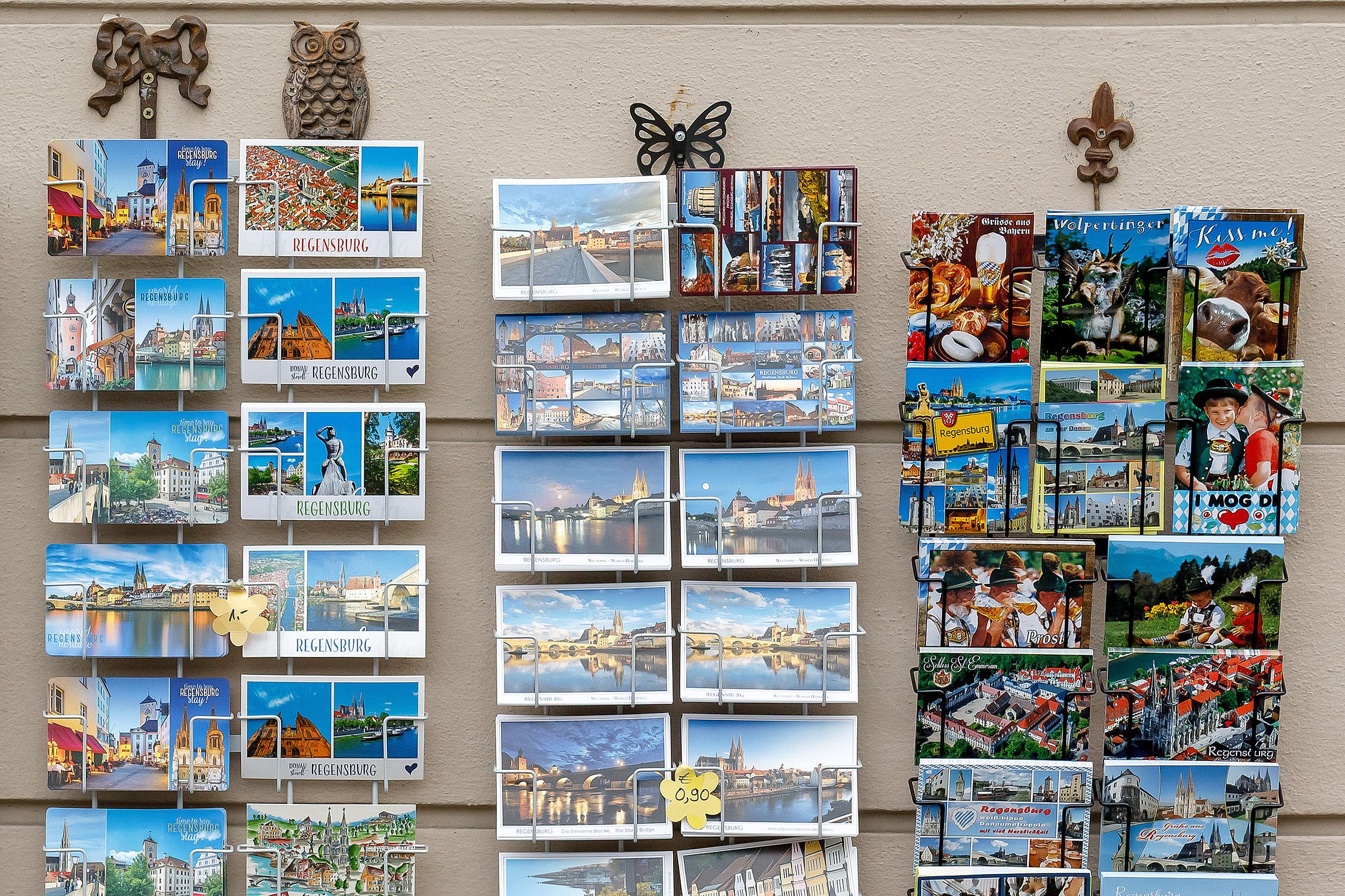 Fotografie: Drei Aufsteller, in denen Ansichtskarten mit unterschiedlichen Regensburg-Motiven präsentiert werden, vor einer grauen Wand
