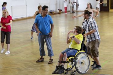 Fotografie - Erwachsene und Mann im Rollstuhl (mit Begleitung) beim gemeinsamen Tanzen