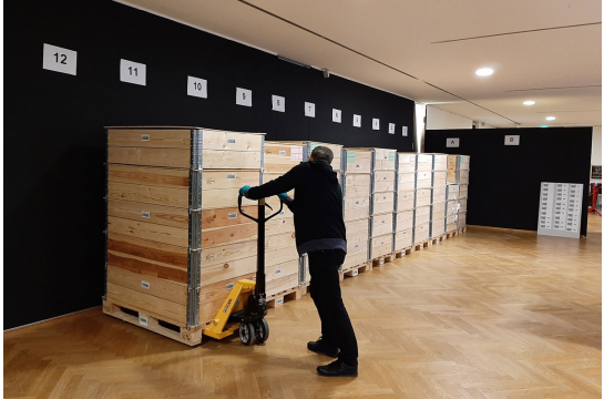 Fotografie: Ein Mann mit Gabelstapler platziert große Kisten vor Nummern an Wänden.