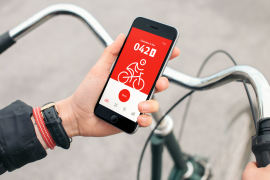 Fotografie: Fahrradlenker im Bildhintergrund, Hand mit Smartphone im Bildvordergrund. Auf dem Smartphone ist die DB Rad+ App geöffnet. 