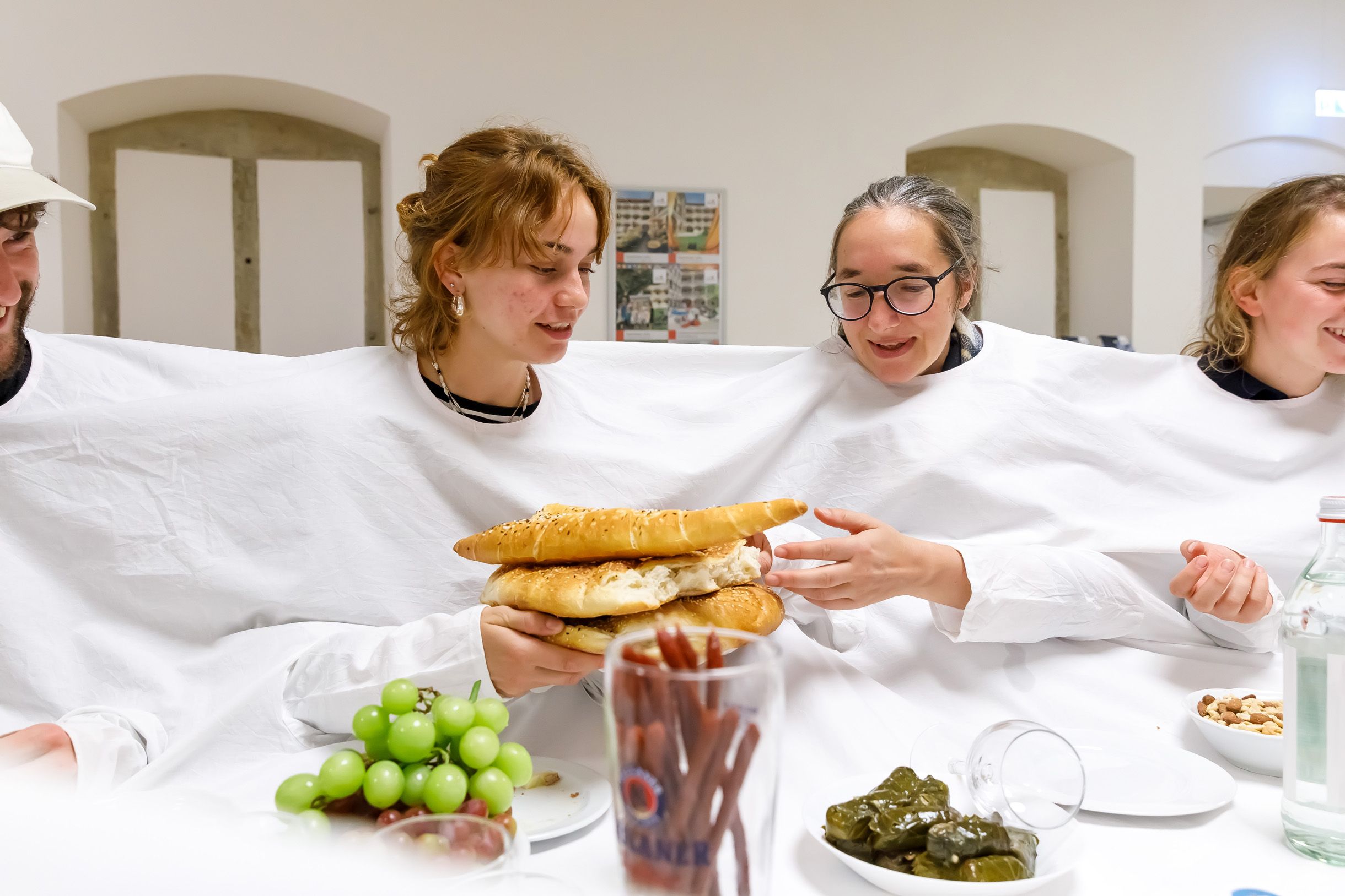 Fotografie: Vier Personen stecken in einem großen weißen Laken mit Öffnungen für Hände und Köpfe und reichen einen Teller mit Fladenbrot durch.
