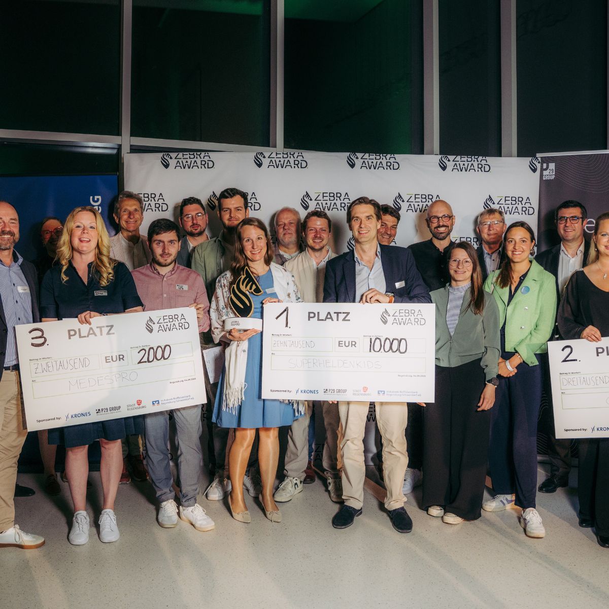 Eine Menschentraube steht vor einer Leinwand, auf dem das Motiv eines Zebras zu sehen ist sowie der Spruch "Zebra Award" drauf gedruckt wurde. Die Menschen in der ersten Reihe halten Preise von 2.000€, 3.000€ und 10.000€ in den Händen.