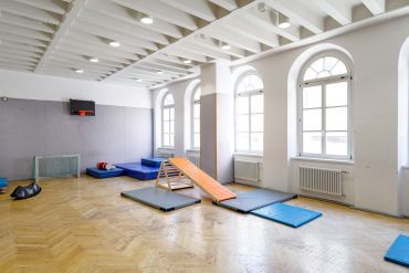 Turnhalle mit Parkettboden und gepolsterter Wand. An der Wand hängt ein Basketballkorb und es liegen verschiedene Matten im Raum verteilt. Auch eine Rollenrutsche und Fußballtore sind zu sehen