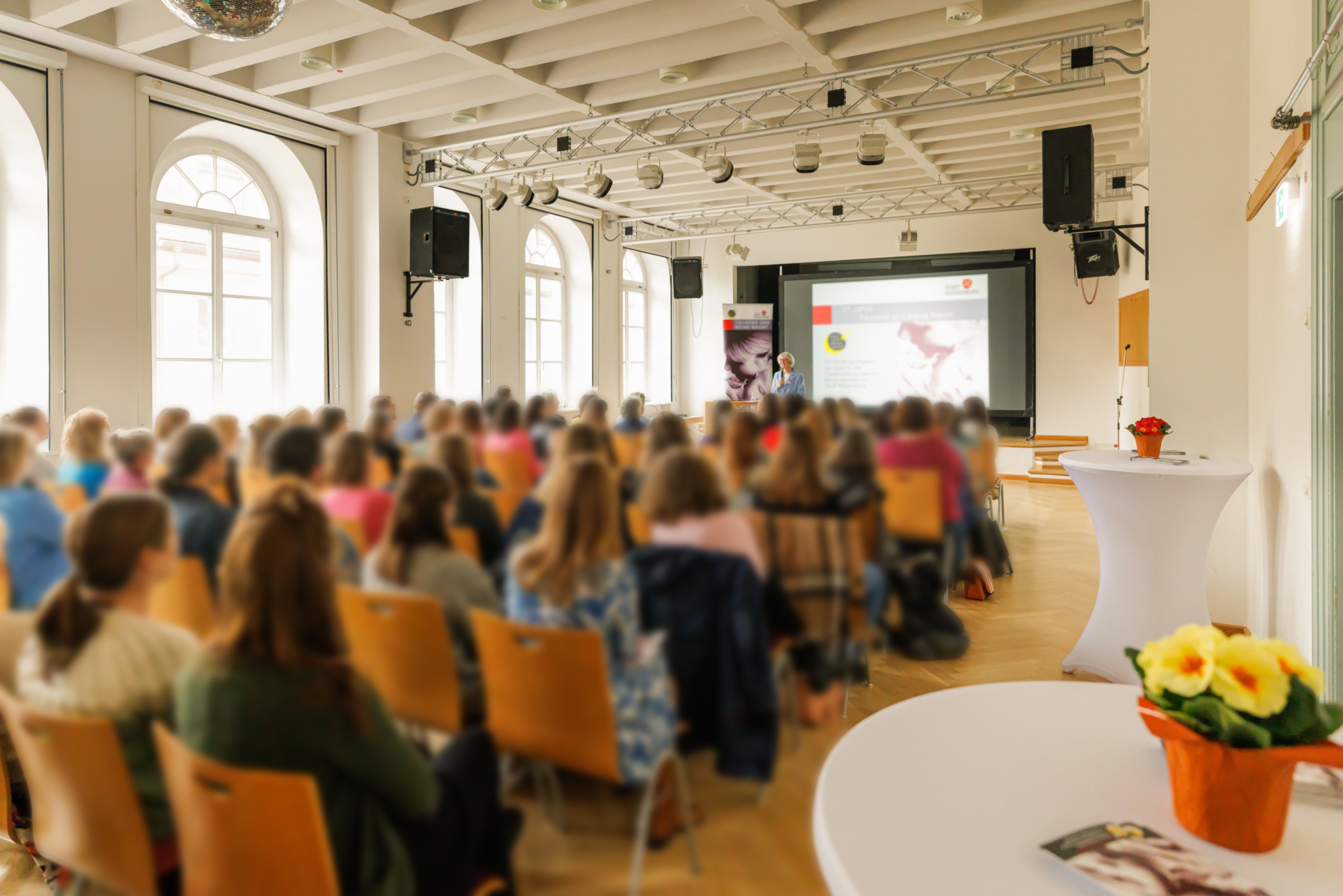großer Saal mit Reihenbestuhlung und ca. 70 sitzenden Gästen. Auf der Bühne steht Leinwand mit laufender Präsentation. Stehtisch mit Blumen im Vordergrund. 