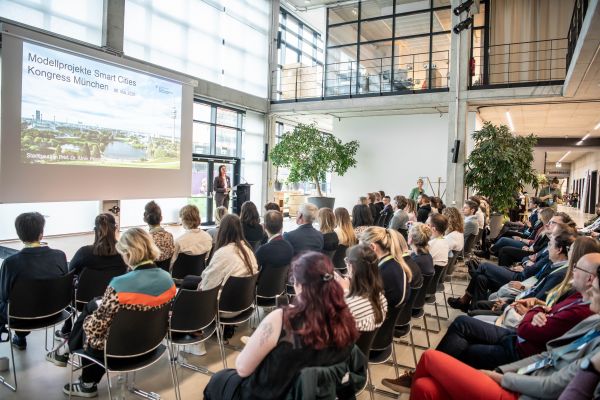 Auf dem Bild erkennt man einen großen Saal, indem man ein großes, sitzendes Publikum erkennt, das auf eine Leinwand schaut. Auf der Leinwand ist die Start-Folie des Kongresses zu erkennen.