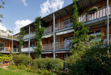 Das Bild zeigt ein Holzgebäude mit einem umlaufenden Balkon aus Metall. Der Garten vor dem Gebäude ist stark bewachsen. An den Halterungen des Balkons wachsen Pflanzen empor.