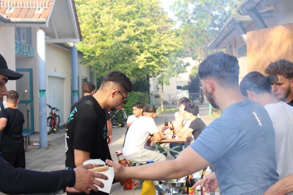 Eindrücke vom Sommerfest des Jugendzentrum Arena. Viele Jugendliche sind zu sehen. Im Vordergrund überreicht ein junger Mann einem anderen einen Burger und im Hintergrund sitzen mehrere junge Männer an einem Tisch und stoßen mit Spezi-Flaschen an