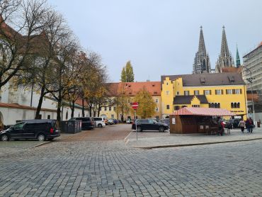 Zu sehen ist ein mit Kopfsteinplaster gepflasterter Platz. Im Hintergrund sind zwei Kirchtürme zu sehen, die halb von einem gelben Haus verdeckt werden. Vor diesem Haus, mittig auf dem Platz gelegen, steht eine Bude aus Holz mit einer rot-weißen Markise. Links im Bild ist eine Straße zu sehen. An dieser ziehen sich Häuser entlang, davor verläuft eine Baumreihe. Zwischen der Baumreihe und der Straße sind Fahrzeuge geparkt.