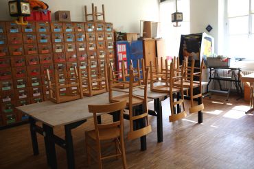 Werkraum mit stabilen Tischen und diversen Holzstühlen. Von der Decke hängen einzelne Steckdosen Verteilerdosen für Werkzeug. Im Hintergrund steht ein Apothekerschrank mit sehr vielen bunt beschrifteten Schubfächern