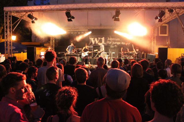 Ein Auftritt der Band "Containerhead" am Regensburger Bismarckplatz mit Blick aus dem Publikum. Im Bühnenhintergrund ein Banner mit dem Logo des W1.